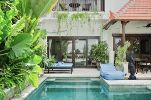 A pool area with a lounge chair and a statue, surrounded by lush greenery and a building with a tiled roof.
