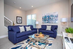 A living room with two blue sofas, a glass coffee table, and a modern abstract painting on the wall. - 4 Suites Villa 5 min to Disney (Kissimmee)