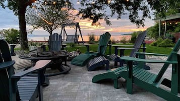 Relax in the outdoor seating area with Adirondack chairs and a fire pit, overlooking a serene body of water.