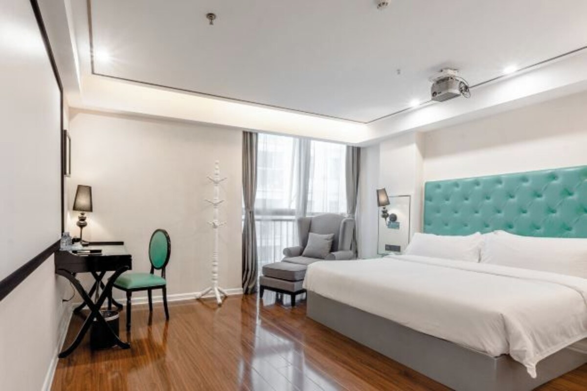 A hotel room with a large bed, a desk with a chair, a floor lamp, and a window with curtains.