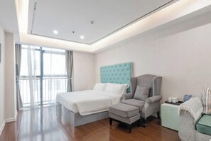 Quarto de hotel moderno com uma cama grande, uma poltrona cinza, uma pequena mesa de apoio e vista para o exterior por uma grande janela.