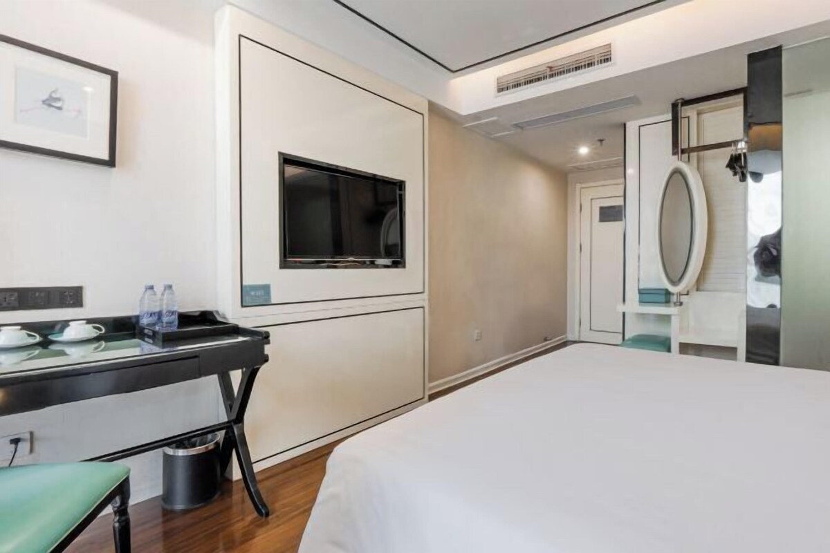 Quarto de hotel com cama, escrivaninha com bandeja, televisão, espelho e porta.