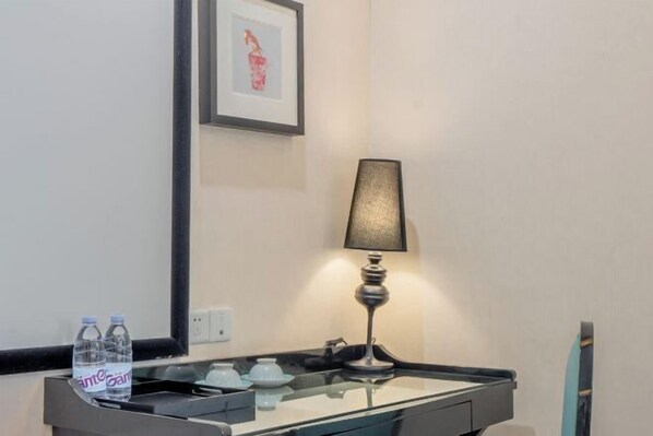 Quarto de hotel com uma secretária de vidro, uma lâmpada, duas garrafas de água e um quadro na parede.