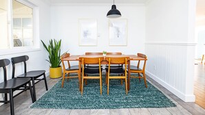 Et spiseområde med et træbord og stole, en sort hængende lampe, billeder i rammer på væggen og en potteplante.