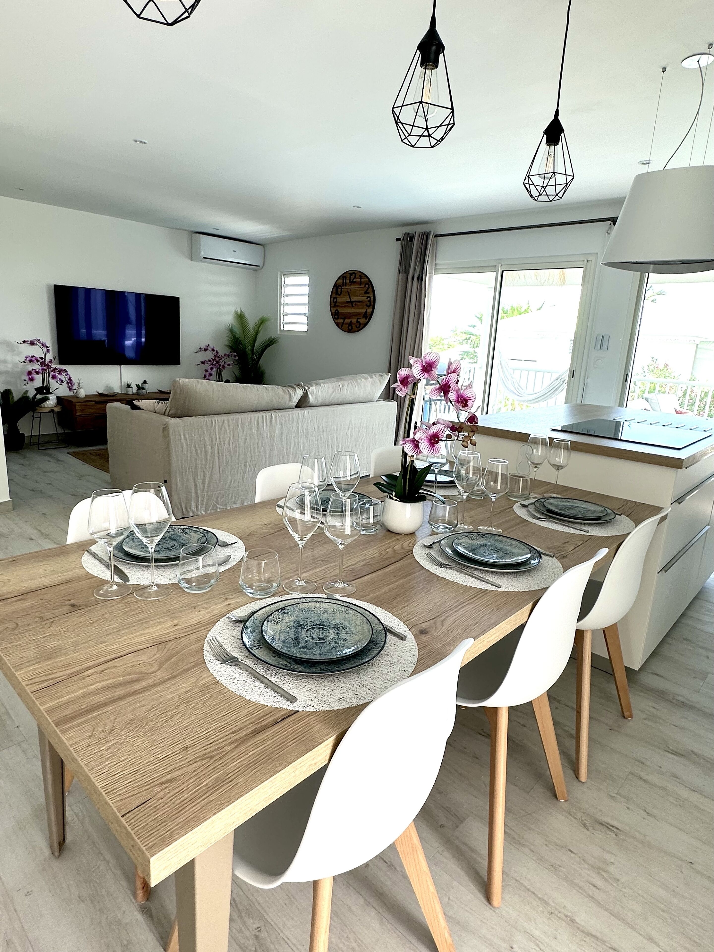 A dining area with a wooden table set for six, white chairs, and a TV in the background.