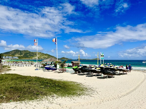 A beach with umbrellas, a water sports area, and a mountain in the background. - Orient Bay, Appartement Luxueux à 3 Minutes de la Plage! (Collectivité de Saint-Martin)
