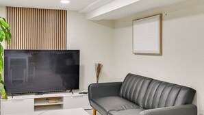 A modern living room with a flat-screen TV on a white entertainment unit, a grey leather sofa, and a wooden slat wall panel. - Lovely Elmont Apartment - Walk to Bus Stop! (Elmont)