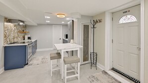 A modern kitchen with a white countertop and blue cabinets, a dining area with a white table and two chairs, and a door with a "No Smoking" sign. - Lovely Elmont Apartment - Walk to Bus Stop! (Elmont)