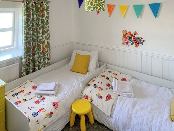 A bedroom with two beds, a yellow stool, a window with curtains, and wall decorations including bunting and a cartoon character.