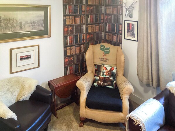 A cozy room with a leather sofa, a plush armchair with a print, a bookshelf, and framed pictures on the walls.