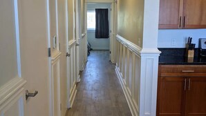 Un pasillo con piso de madera, paneles de madera blancos y una zona de cocina con gabinetes de madera y encimera negra.