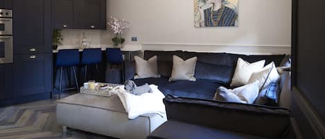A modern living room with a dark-colored sofa, a coffee table, and a kitchen area with dark cabinets and a white countertop.