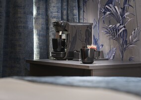 Una cafetera, una taza y un pequeño recipiente sobre una mesa en una habitación con un papel tapiz estampado.