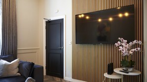 A modern living room with a black sofa, a flat-screen TV mounted on a wooden panel wall, and a small table with a potted orchid. - Home at Tancred Road  (Liverpool)