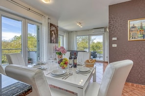 A dining area with a white table set for four, a fruit bowl, wine bottles, and a basket of bread. - Sea Breeze Residence Mariella (Umag)
