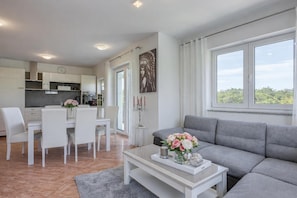 A modern living room with a grey sofa, a dining area with white chairs, and a kitchen in the background. - Sea Breeze Residence Mariella (Umag)