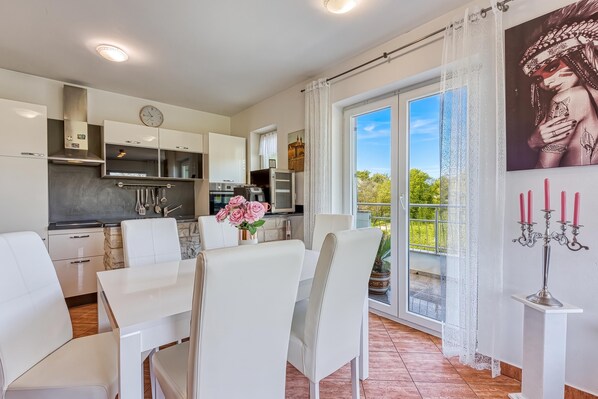 A modern kitchen with white cabinets, a dining area with white chairs, and a balcony with a view of greenery.