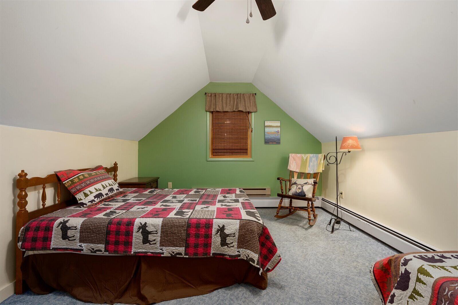 Un dormitorio con una pared verde de acento, una cama de madera con colcha, un sillón mecedor y una lámpara de pie.