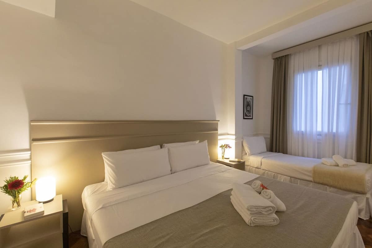 A hotel room with a large bed, a smaller bed, a nightstand with a lamp, a vase with flowers, and a window with curtains.