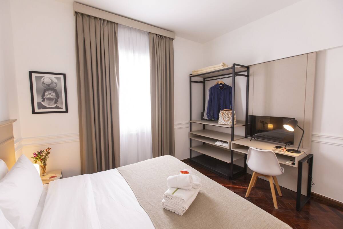 A hotel room with a bed, a desk with a chair, a wardrobe, and a large window with curtains.