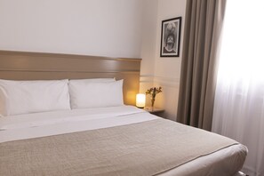 Una cama bien tendida con sábanas blancas, una lámpara de noche y un cuadro en la pared.