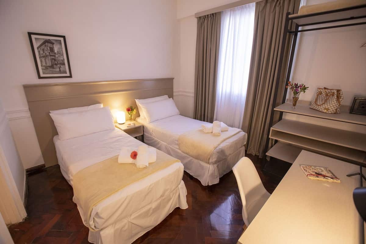 A hotel room with two beds, a nightstand with a lamp, a desk, and a chair.