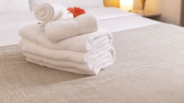 Una cama bien hecha con sábanas blancas y una toalla enrollada encima, una lámpara de noche y un cuadro en la pared.