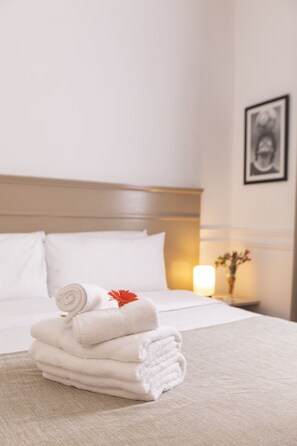Una cama bien hecha con sábanas blancas y una toalla enrollada encima, una lámpara de noche y un cuadro en la pared.
