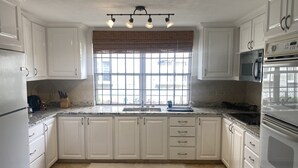 Une cuisine avec des meubles blancs, un réfrigérateur en acier inoxydable, un four à micro-ondes et un four traditionnel. Il y a une fenêtre avec des stores et un luminaire au-dessus de l’évier.