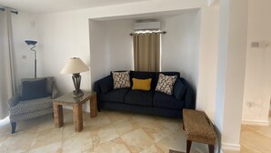Un salon avec un canapé bleu foncé, des coussins à motifs, une petite table avec une lampe et un ventilateur de plafond.