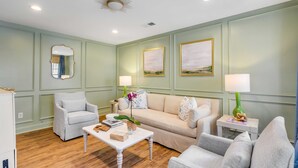 Un salon avec un canapé beige, deux fauteuils, une table basse et une table d’appoint avec une lampe. Des tableaux encadrés ornent le mur, et un miroir est placé au-dessus du canapé.