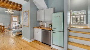Una cocina moderna con armarios blancos, lavavajillas y un refrigerador verde claro. Hay una mesa de comedor de madera con sillas y un sofá en la zona de estar contigua.