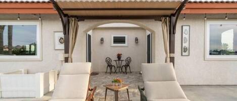 A covered outdoor seating area with two lounge chairs, a small table, and a view of the interior through an archway.