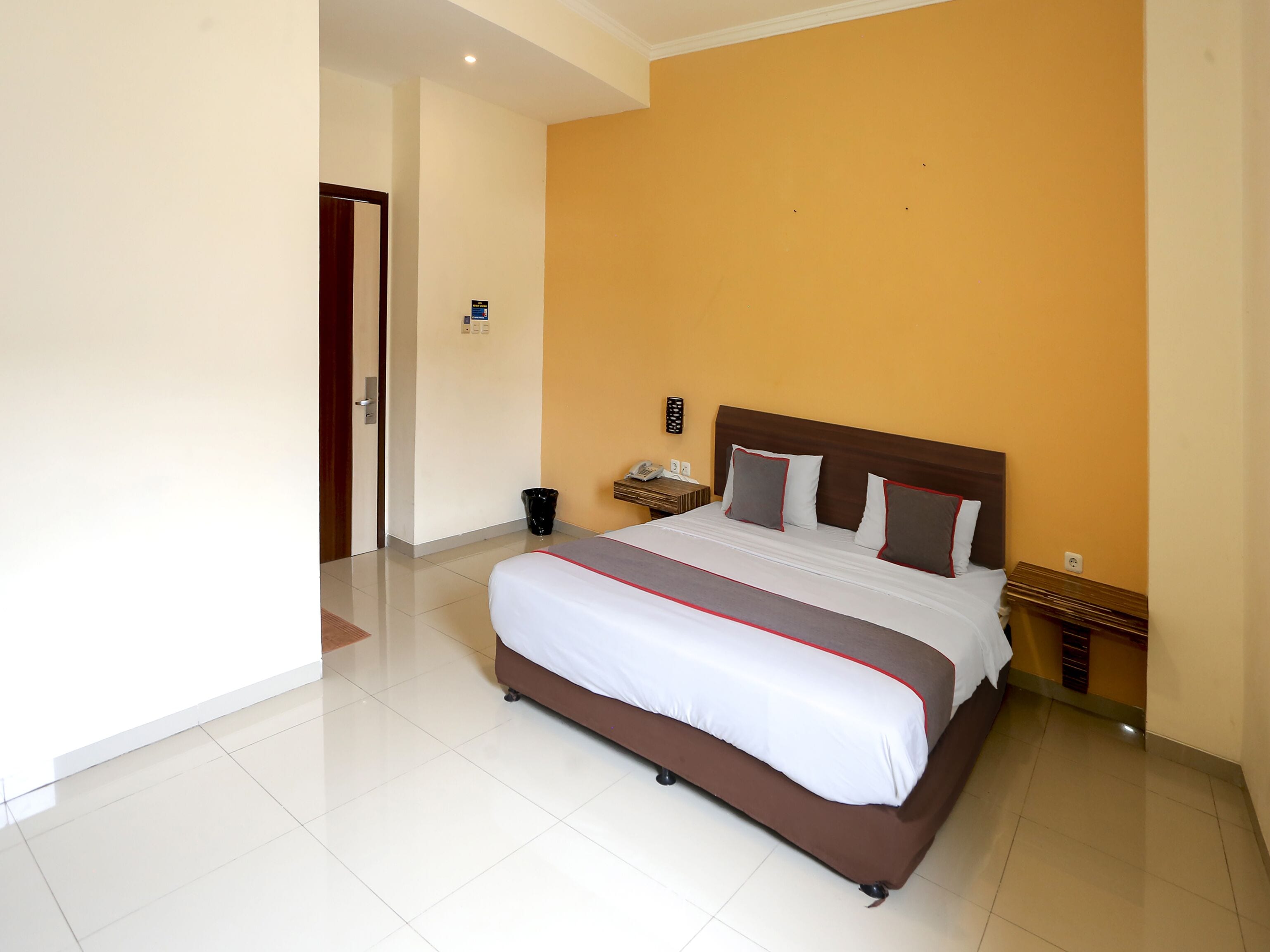 A hotel room with a large bed, a bedside table with a phone, and a wall-mounted control panel.