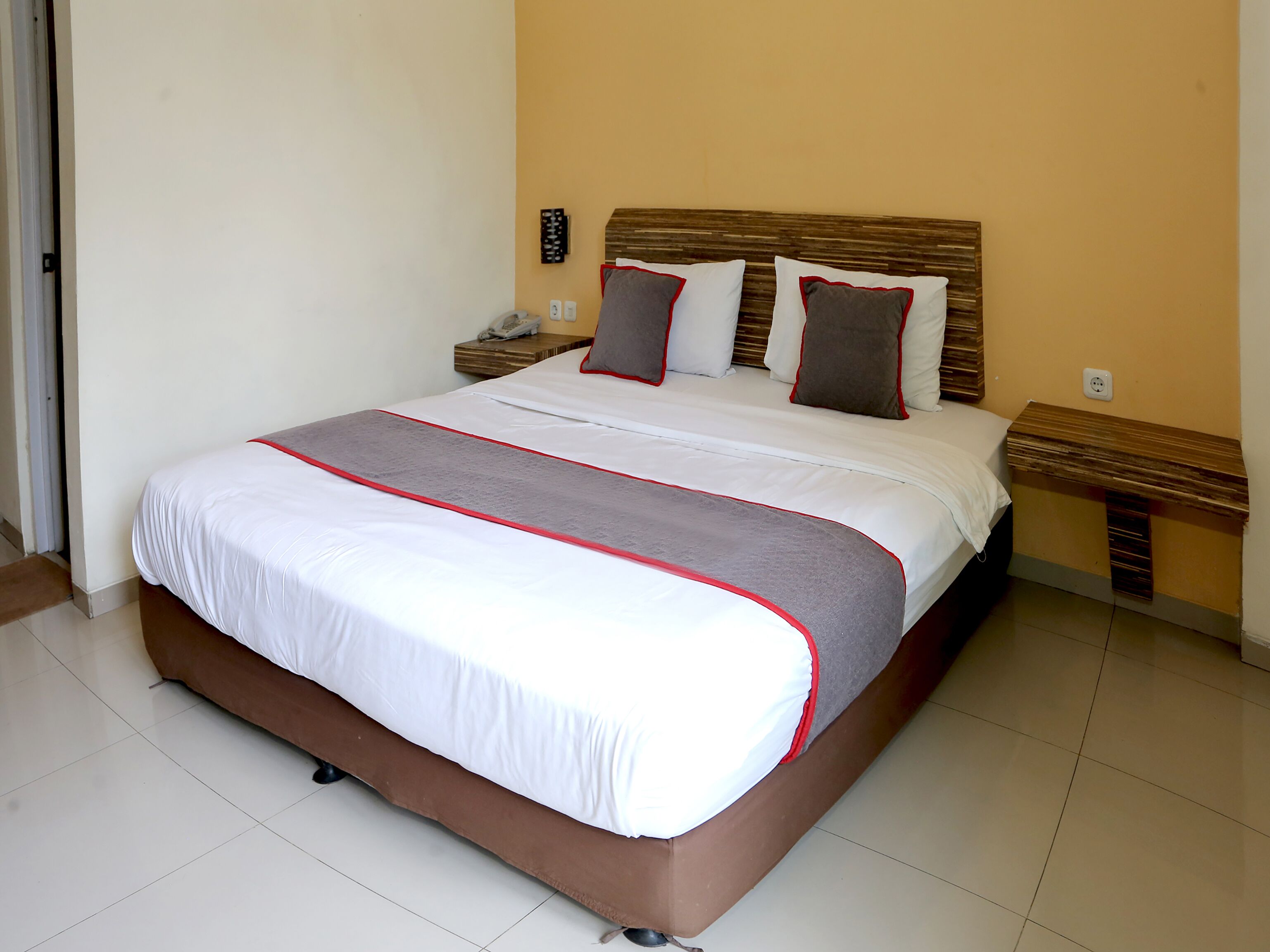 Una cama bien tendida con una colcha roja y blanca, dos mesitas de noche con lámparas, y un aplique de pared.