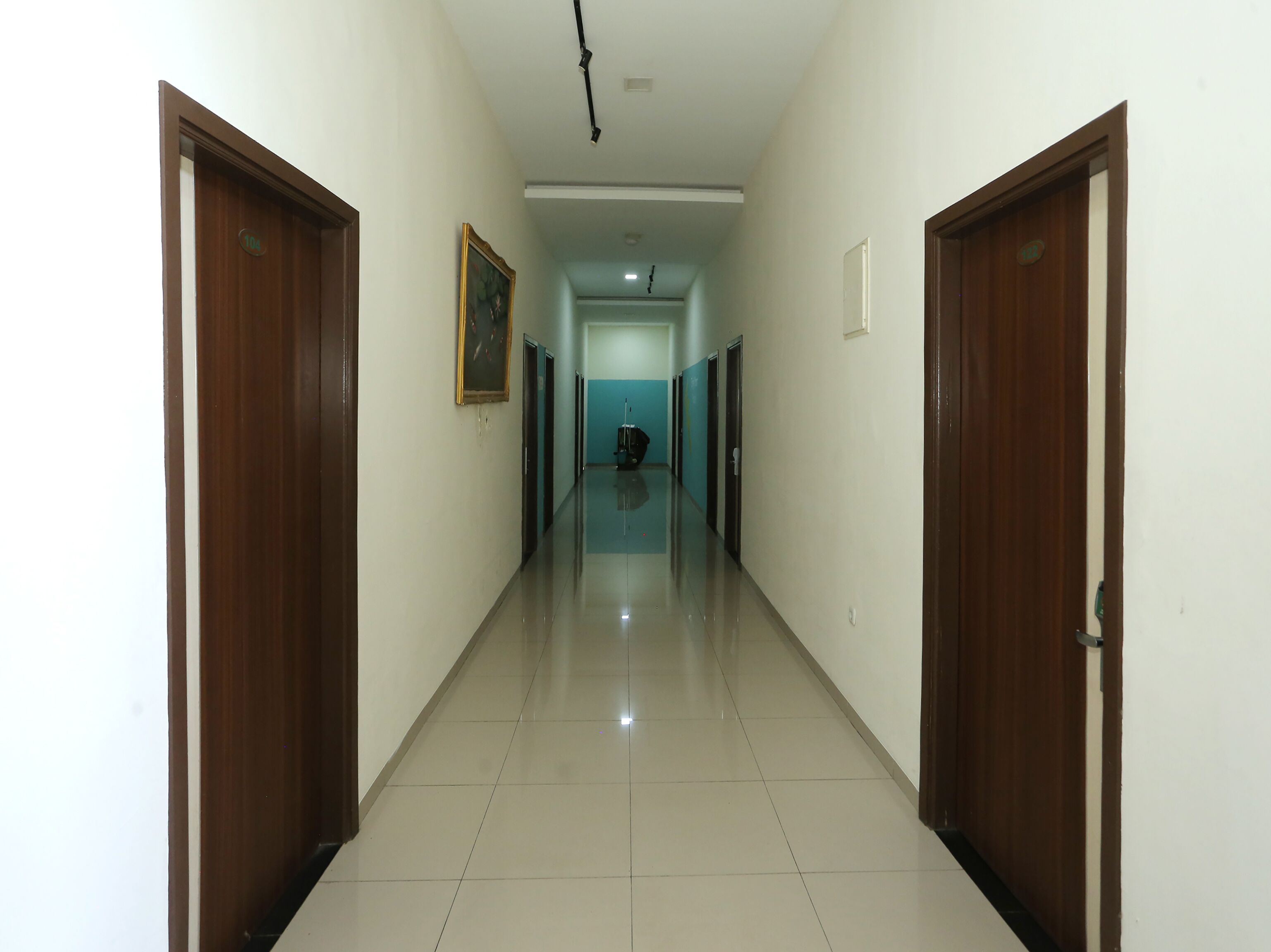 A long, narrow hallway with wooden doors on either side, leading to a room with a blue wall and a ceiling light fixture.