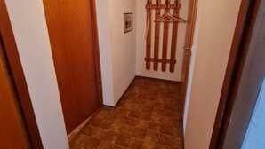 Ein schmaler Flur mit einer Holztür links, einem gefliesten Boden und einem Kleiderständer mit Kleiderbügeln rechts.