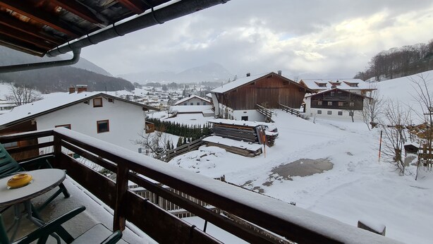 Ein verschneites Dorf mit traditionellen Holzhäusern, einem Holzbalkon und Blick auf ferne Berge.