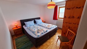 Ein Schlafzimmer mit einem Bett, zwei Nachttischen, einem Sessel, einem Fenster mit Vorhängen und einer Pendelleuchte.