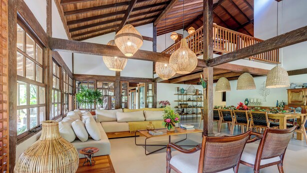 A spacious living area with a high ceiling, wooden beams, and large windows. There is a sofa, a coffee table, and wicker chairs. - Casa OluOlu Itacaré (Itacaré)