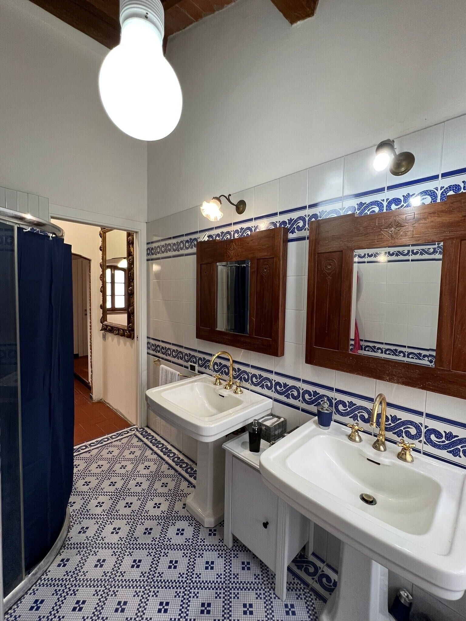 Casa de banho com duas pias, chuveiro e espelho com moldura de madeira. O chão tem um padrão de azulejos.