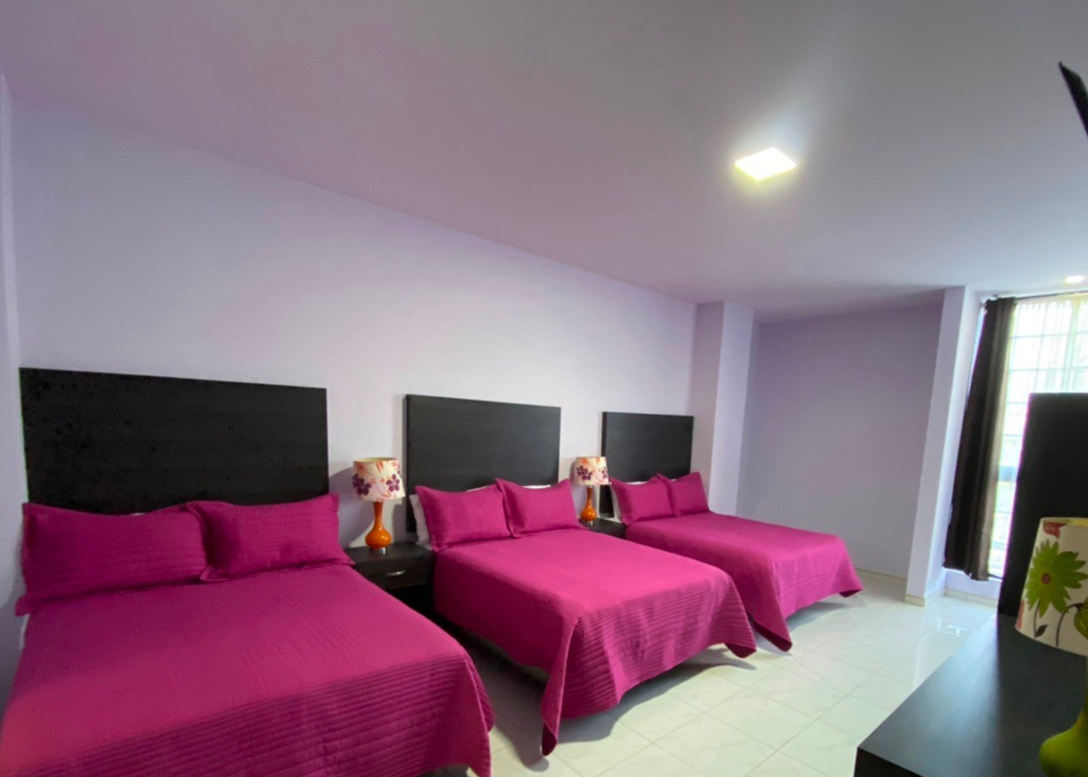 A hotel room with three beds, each with a black headboard and pink bedding. There are bedside tables with lamps and a large window with curtains.