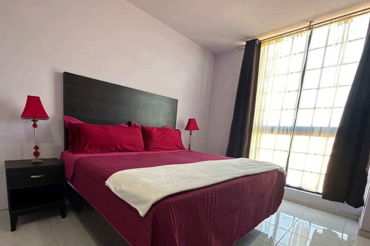 A neatly made bed with red bedding and a white throw, a black nightstand with a red lamp, and a window with curtains.