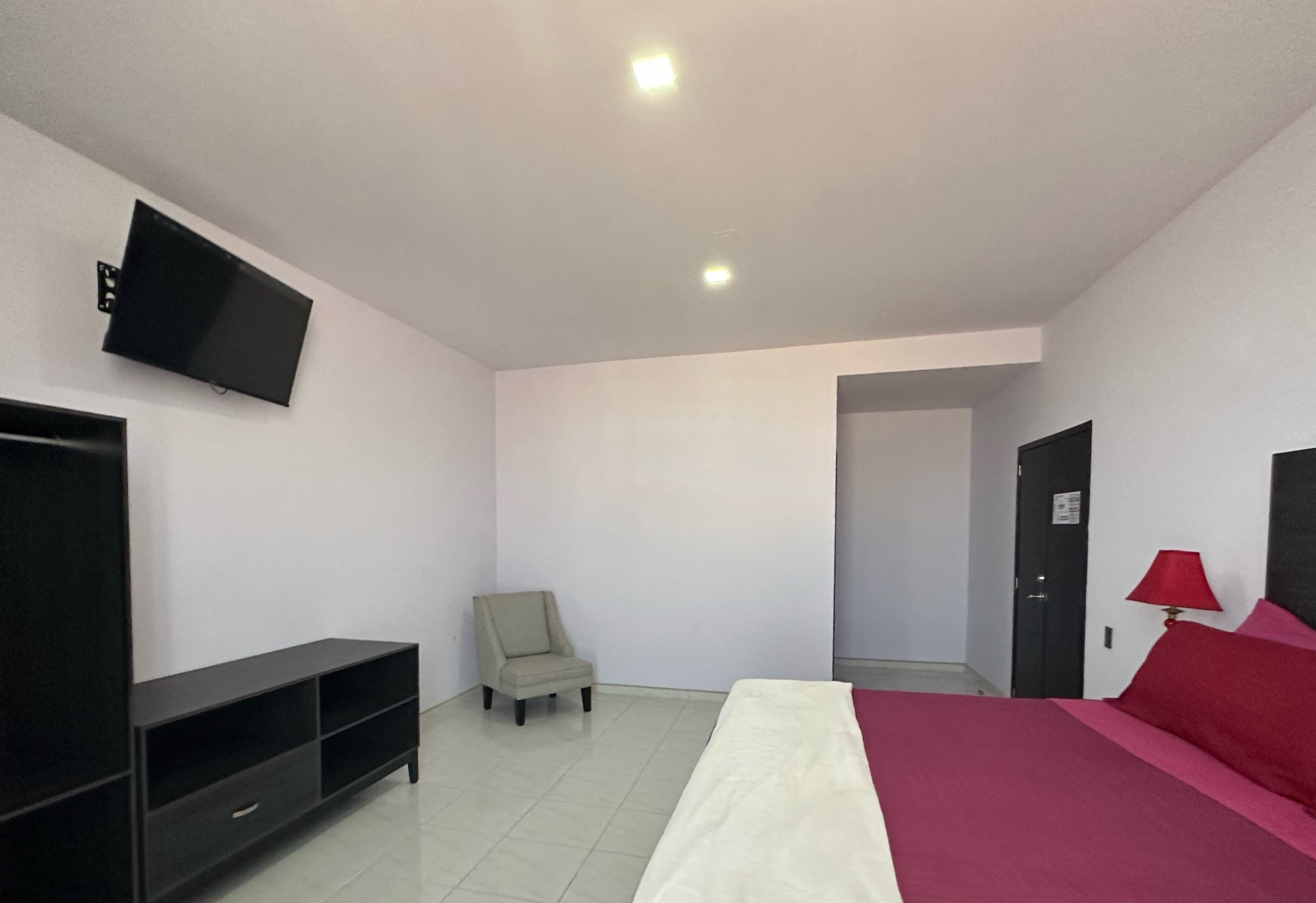 A bedroom with a bed, a TV mounted on the wall, a chair, and a door leading to another room.