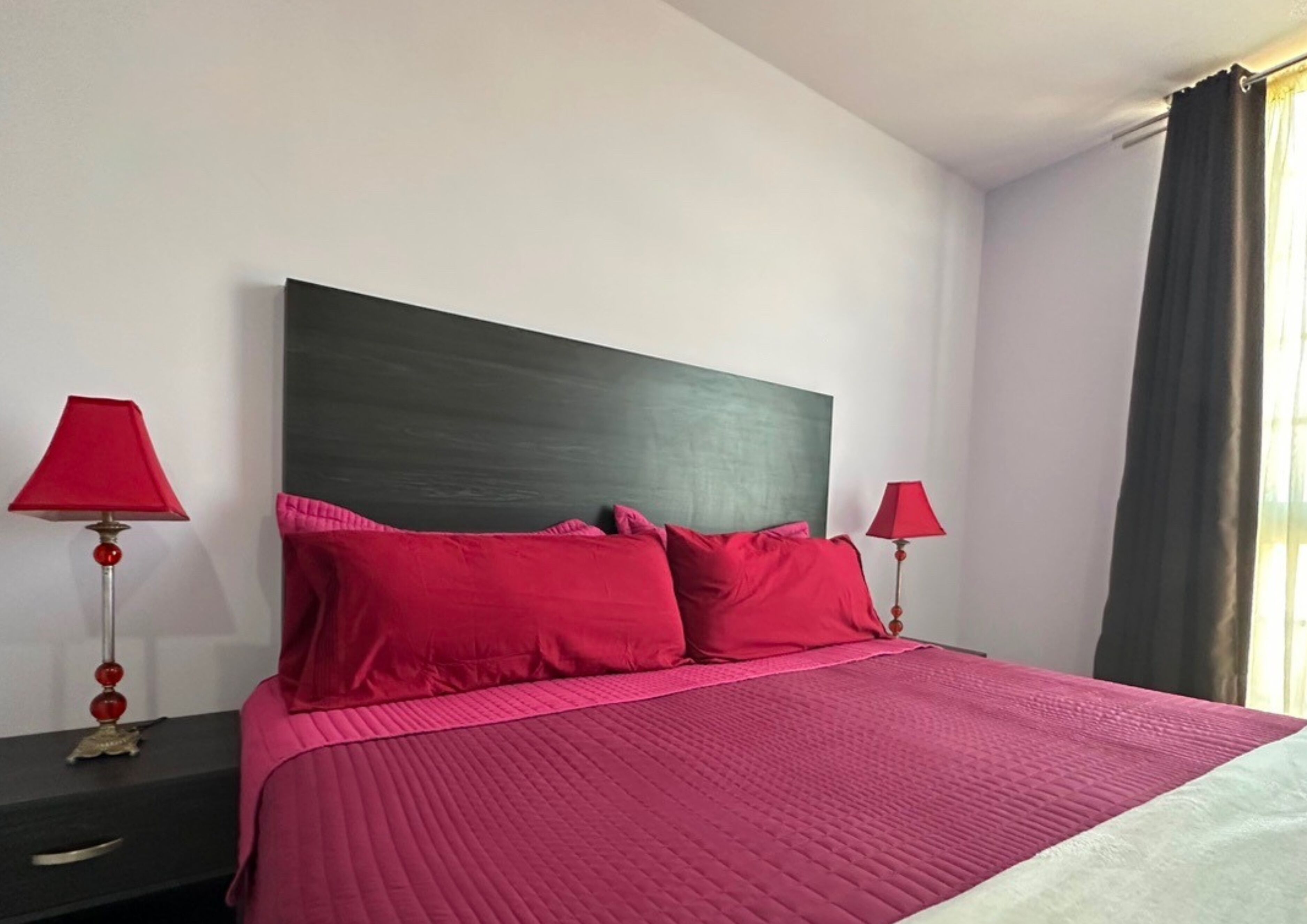A bed with a dark headboard, two red lamps, and a window with curtains.