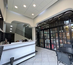 Vestíbulo de hotel con mostrador de recepción, una escalera y un separador de vidrio que ofrece vistas a la calle.