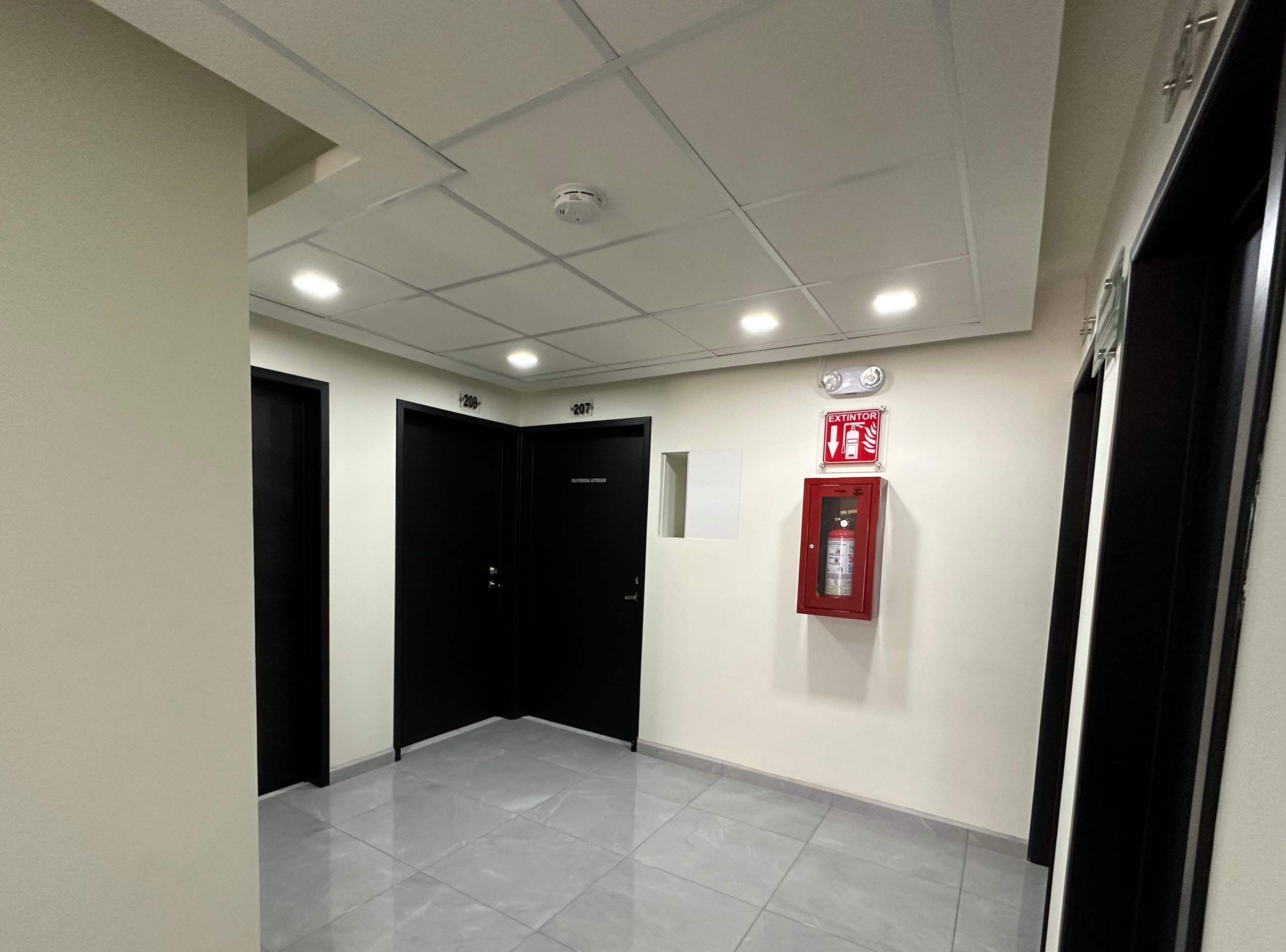Un pasillo con puertas negras, un extintor y un techo con iluminación empotrada.