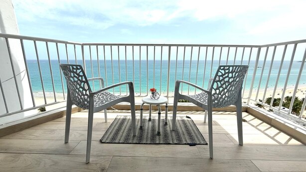 A balcony with two metal chairs and a small table, overlooking a beach with clear blue water and a sandy shore. - Mb17- One Bedroom with Ocean Front Balcony (Carolina)