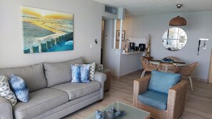 A living room with a grey sofa, wicker chairs, a coffee table, a painting of a beach, and a kitchen area with a round mirror and a hanging light fixture.