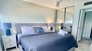 A modern bedroom with a large bed, a nightstand, a mirror, and a door leading to another room. - Mb17- One Bedroom with Ocean Front Balcony (Carolina)